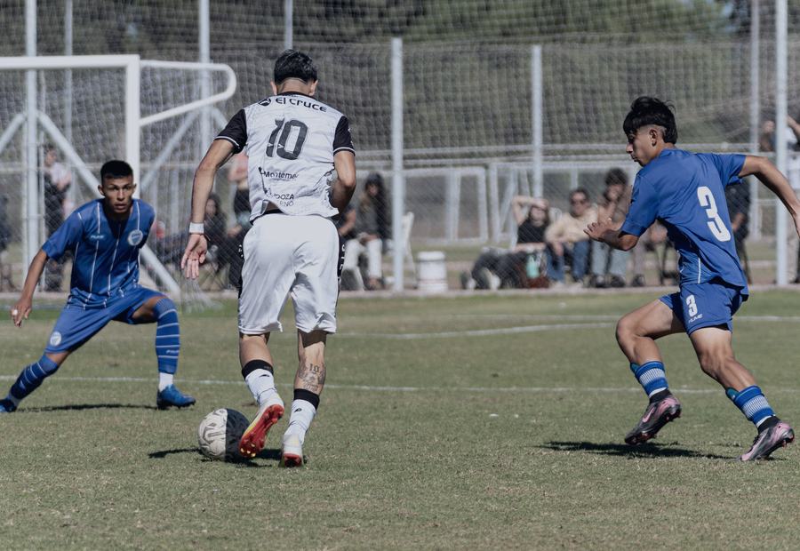 Partido de fútbol formativo en cancha exterior, con público en la línea